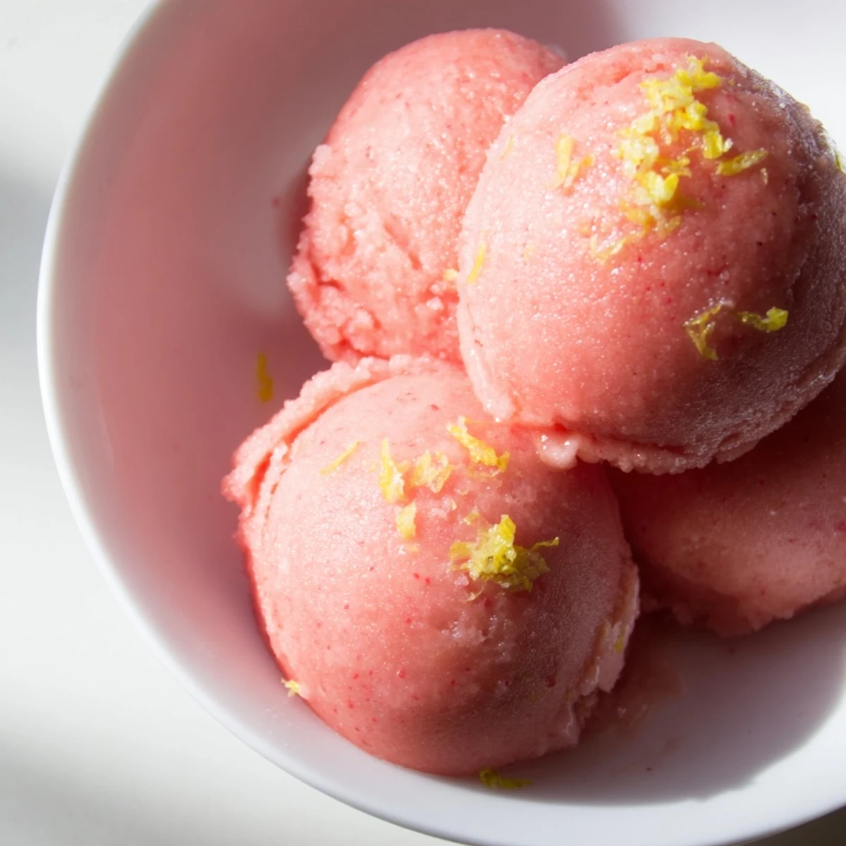 A close-up of vibrant red Strawberry Lemonade Sorbet in a chilled bowl, garnished with a fresh mint leaf and lemon zest.  