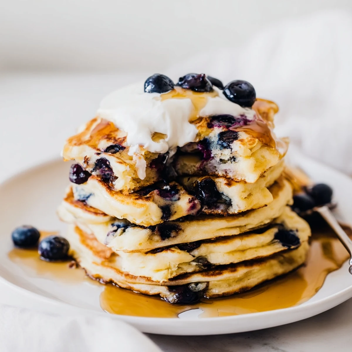 A warm skillet of Fluffy Greek Yogurt Blueberry Pancakes sizzling next to a pitcher of maple syrup and a bowl of blueberries.