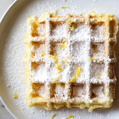 A stack of fluffy Lemon Ricotta Waffles with a dusting of powdered sugar, served alongside a cup of coffee.